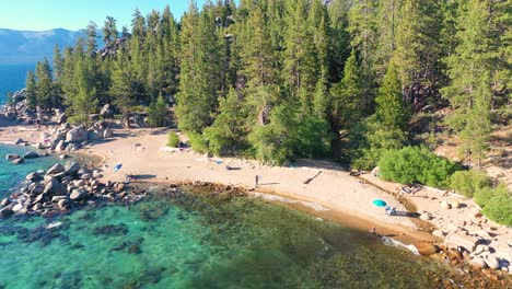 playa pública en el lago tahoe, california con espesos bosques de pinos en el fondo, gente disfrutando del sol, aguas azules claras y grandes rocas - retirada aérea de drones