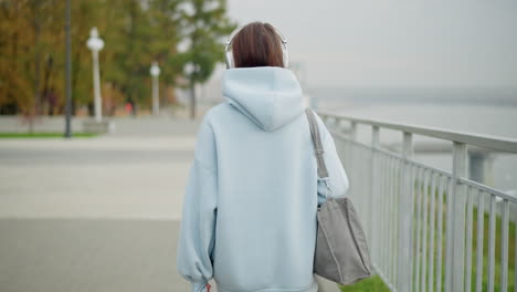 Back view of woman in blue sweater and headphones holding bag, walking slowly along iron rail, serene outdoor atmosphere, peaceful morning walk, urban setting, casual lifestyle