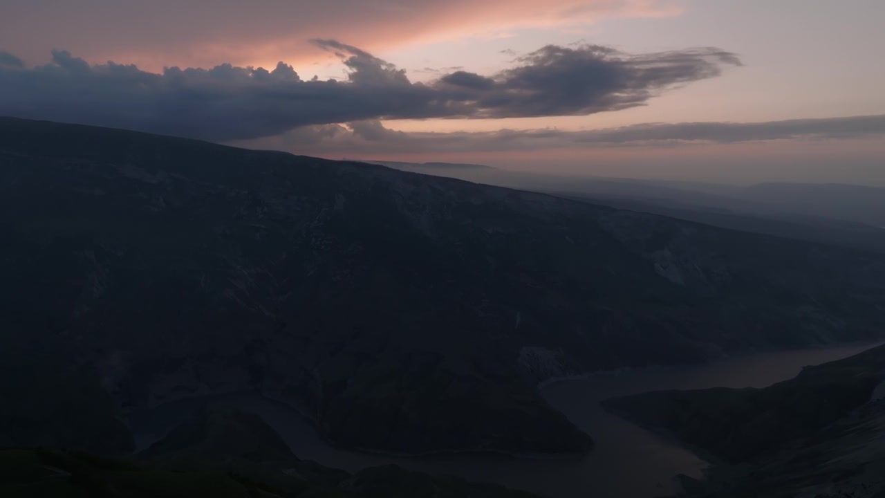 Aerial footage of Sulak Canyon with a winding river between dark mountain ridges at sunset, dramatic clouds and soft haze over Dagestan, Russia