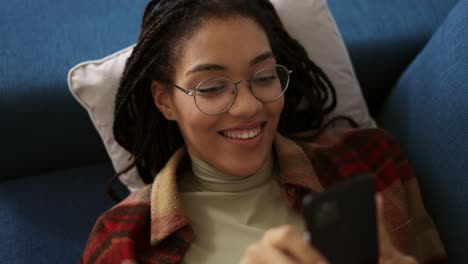 happy african american teen relaxing on sofa at home with smartphone