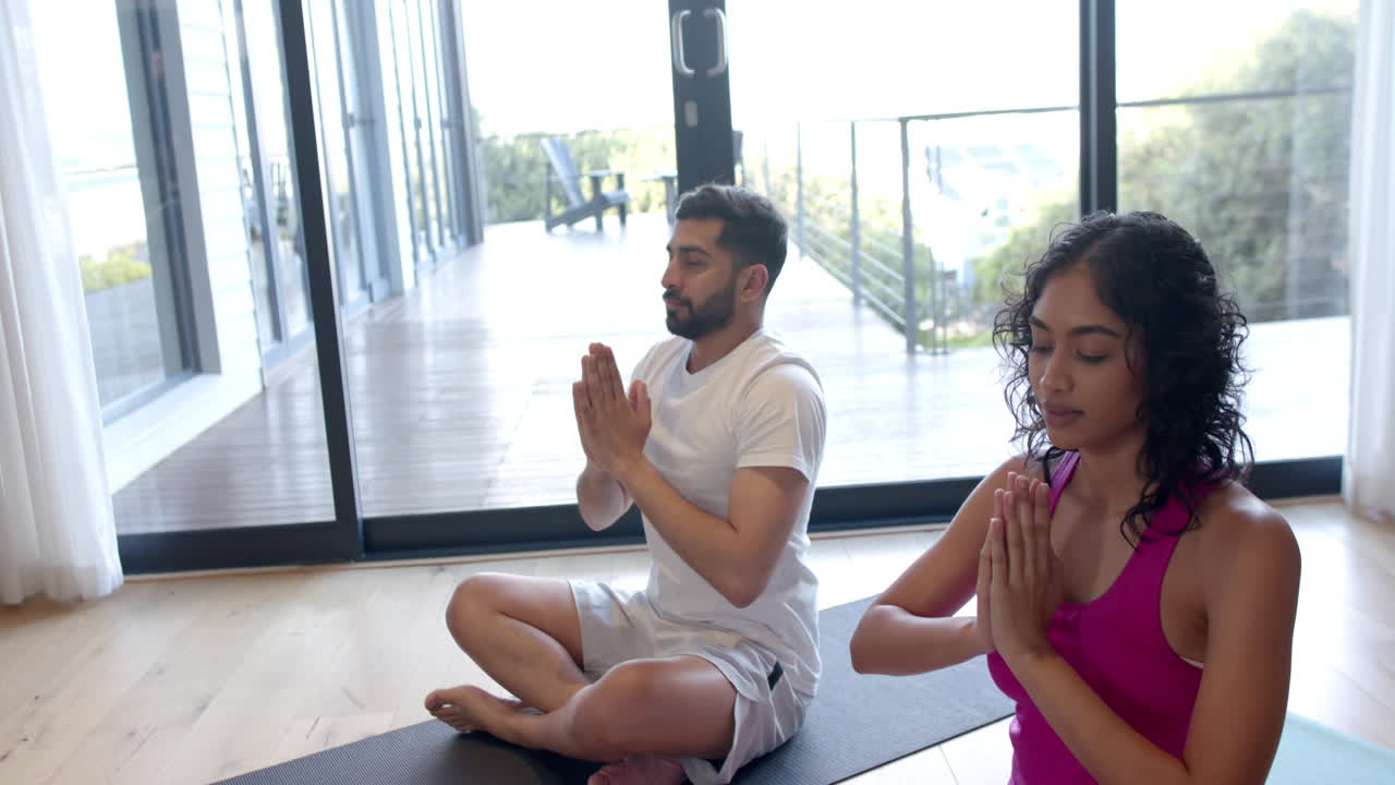 pareja biracial haciendo yoga y meditando en casa, en cámara lenta