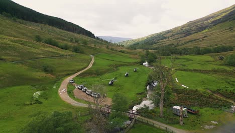 Aerial-Pullback-of-Helicopters-Landed-in-Lush-Green-Valley-next-to-4x4-Vehicles-Convoy-Surrounded-by-Forest-and-Mountains-in-the-Scottish-Highlands,-Scotland,-United-Kingdom