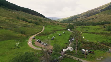 aerial pullback of helicopters landed in lush green valley next to 4x4 vehicles convoy surrounded by forest and mountains in the scottish highlands, scotland, united kingdom