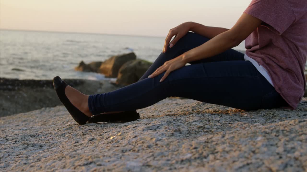 Peinzende vrouw van gemengde afkomst zittend op een rots op het strand tijdens zonsondergang 4k