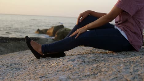 Mujer-Pensativa-De-Raza-Mixta-Sentada-Sobre-Una-Roca-En-La-Playa-Durante-La-Puesta-De-Sol-4k