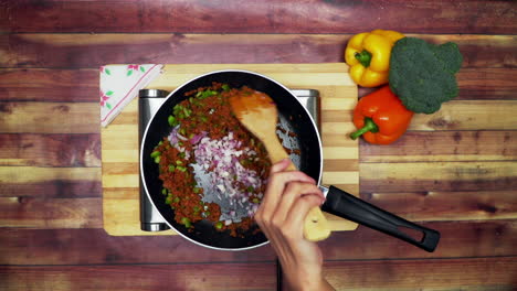 una vista superior de la adición de cebolla a la sartén, colocada sobre una estufa, dos grandes pimientos amarillos y rojos y un brócoli verde sobre la mesa