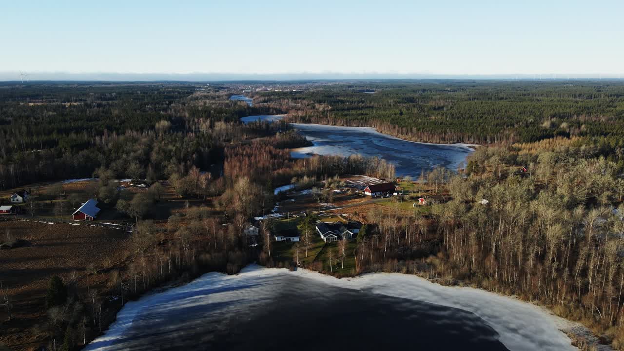 Drone flying towards beautiful vacation homes at the edge of a frozen lake on a sunny day in rural Sweden