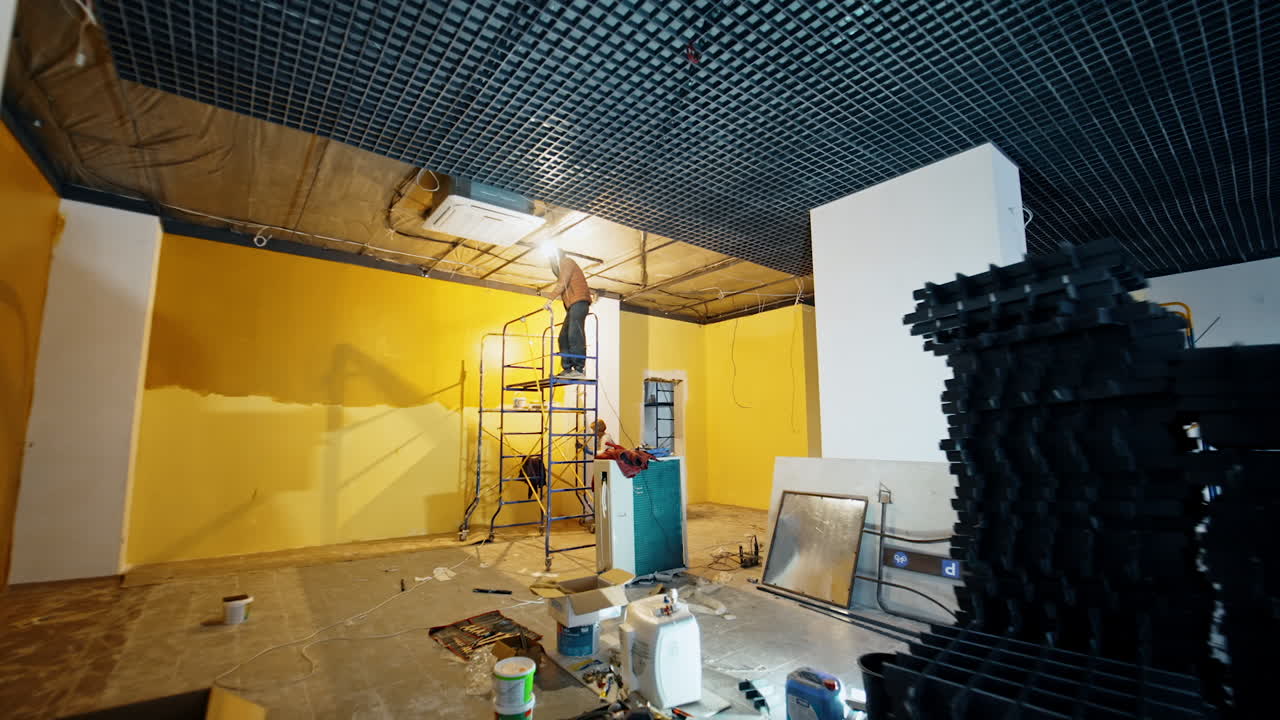 Man stands on the scaffold tower indoors installing light and decor on the ceiling of the room. Overlook of the spacious room with repair work being held by the building team.