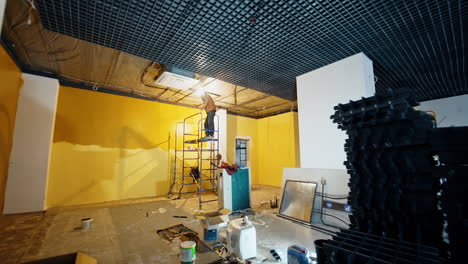 man stands on the scaffold tower indoors installing light and decor on the ceiling of the room. overlook of the spacious room with repair work being held by the building team.
