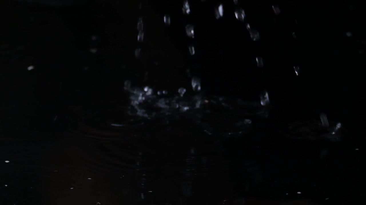 Closeup of water droplets falling into shallow pool puddle of water on clean dark background