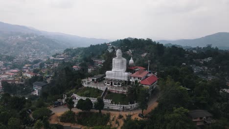 beautiful white bahirawakanda vihara buddha statue on top of a green hill in sri lanka with the large city of kandy in the background between the high mountains on a cloudy day. drone panning shot