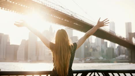 back view of girl with hair blowing in the wind standing at river embankment, arms open near brooklyn bridge new york