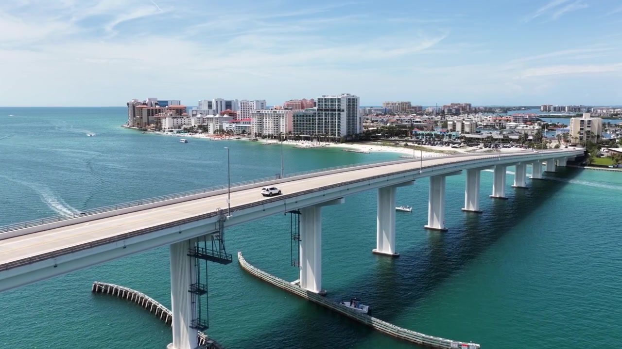Lungo ponte rialzato si estende su acque turchesi verso lo skyline costiero, piccole imbarcazioni punteggiano la baia calma sotto un cielo estivo limpido e azzurro. Clearwater, Florida, USA. Ripresa aerea con drone in avvicinamento