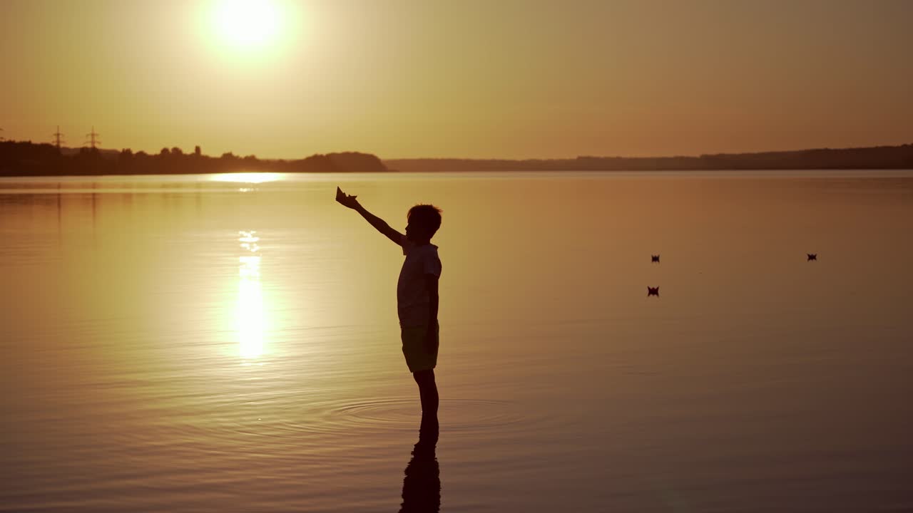 Child in water at sunset. Silhouette of a boy putting paper boat on water in the evening. Kid holding origami ship on the lake.