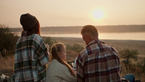 Close-up-shot-of-a-happy-brunette-girl-in-a-green-checkered-shirt-sitting-on-a-mat-with-her-middle-aged-man-husband-and-little-daughter,-they-look-at-the-sunset-and-enjoy-the-view-from-the-hill-during-their-picnic-outside-the-city