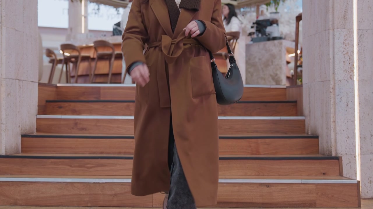Donna che scende scale di legno con il cappotto che ondeggia, borsa di pelle in mano, passo deciso su pavimento in marmo lucido, sfondo di caffè festivo, primo piano di orlo e scarpe, movimento di tracking cinematografico