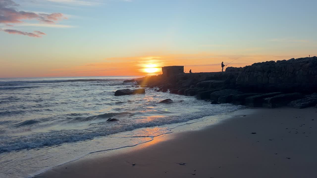 Statisk strandbilde av solnedgang over en steinmolo med fiskersilhuett og Atlanterhavsbølger i Barbate Cadiz.