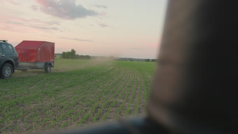 pickup truck driving across green farmland leaving dust trail under pastel evening sky with distant trees and faint clouds on horizon captured from low angle