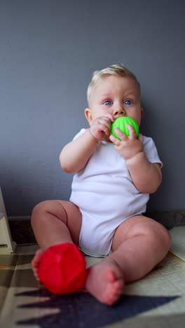 adorable little kid wearing white bodysuit sits at home near the wall. lovely baby holds the green ball at his mouth and red ball at his feet. vertical video.