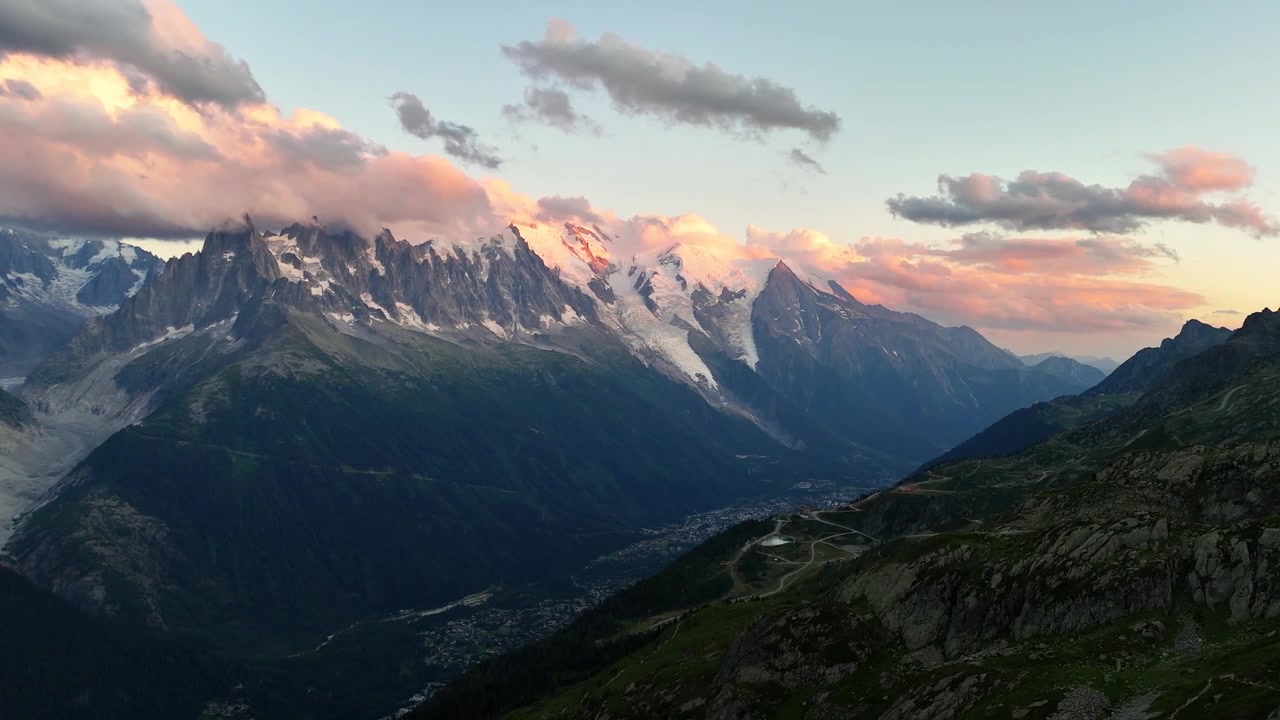 Une vue panoramique s'étend sur la vallée de Chamonix en France vers les sommets déchiquetés et les glaciers du Mont Blanc, tandis que la lumière rose du coucher de soleil colore la neige, les nuages et les pentes rocheuses au-dessus de la ville sombre en contrebas.