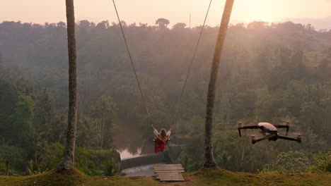 tourist woman swinging over tropical rainforest at sunrise travel girl sitting on swing with scenic view enjoying freedom on vacation having fun holiday with drone flying