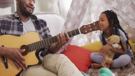 Happy-african-american-father-and-daughter-sitting-in-makeshift-tent,-playing-guitar-and-singing