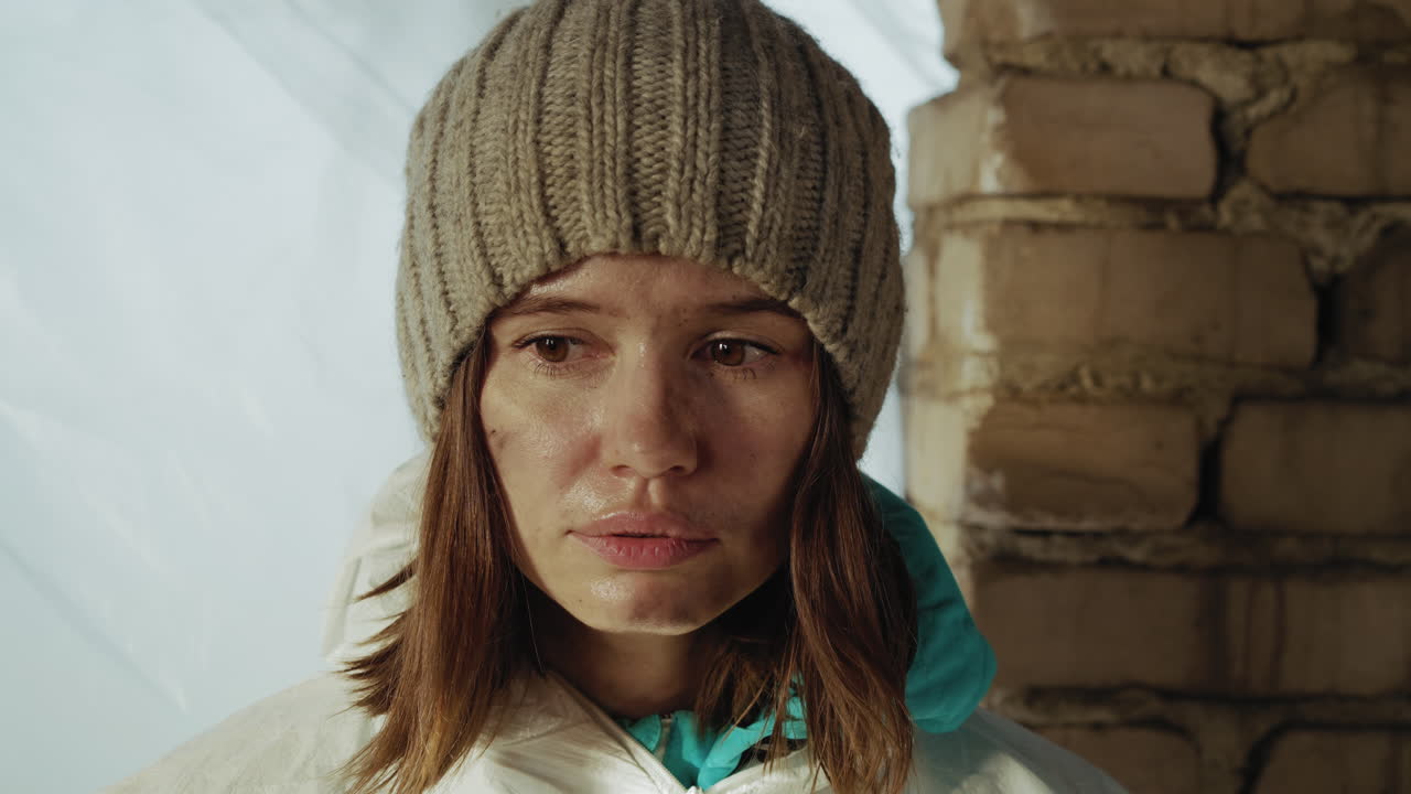Displaced youth wearing knitted beanie and protective suit stands near damaged brick wall with weary smudged face, showing fatigue, confusion, and emotional struggle in aftermath of disaster
