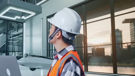 vista lateral de cerca de un ingeniero masculino asiático con casco de seguridad trabajando en una computadora portátil y mirando a su alrededor mientras está de pie con el proyecto de drones