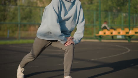 partial view of athlete in blue sweater exercising outdoors with blurred background featuring greenery, bar fence, and seat with another person seated, emphasizing movement and fitness