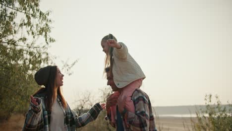 a little blonde girl in a white jacket sits on the shoulders of her dad, the brunette in a brown checkered shirt, and next to her is her mother, the brunette girl in a green checkered shirt, they have fun together, communicate and travel outside the city