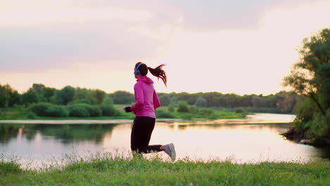 brunette with long hair in headphones runs along the river in the park in the morning at sunrise in the summer in a pink jacket and black pants