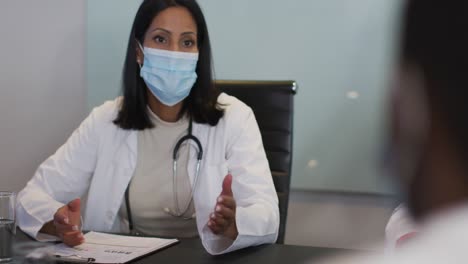 Mixed-race-doctor-sitting-at-table-in-meeting-room-wearing-mask-speaking