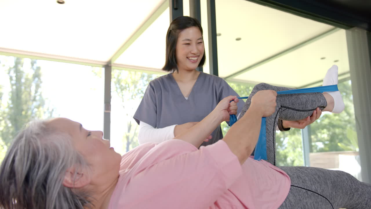 Assisting senior woman with resistance band exercises, physiotherapist smiling in support