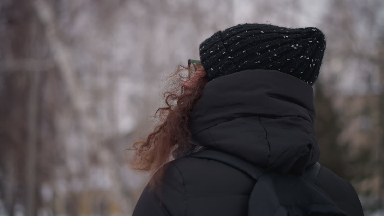 mujer de pie en un parque nevado en invierno, vista lateral, con abrigo negro, gorro, mochila, pelo rizado, soledad, estación fría, aire libre, ambiente gélido, entorno helado, fondo tranquilo, cielo nublado, día apacible