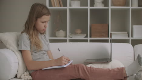 la niña de la escuela está aprendiendo en línea viendo la lección en la pantalla de la computadora portátil y haciendo ejercicios en el cuaderno sentada en el sofá en la sala de estar durante el día