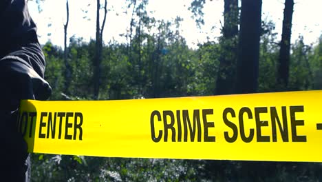 yellow colored crime scene tape being unrolled or unraveled in front of a sunny forest wooded area by an ivestigator who is wearing black nylon gloves. ribbon backlit by sun and is shiny, white sky