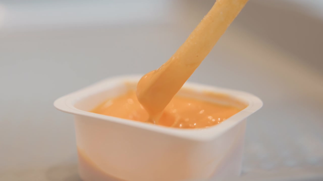 Close view of creamy orange condiment flowing into small single serve plastic cup with visible drip, splash, ripples, and dipping action creating glossy surface motion in food service setting