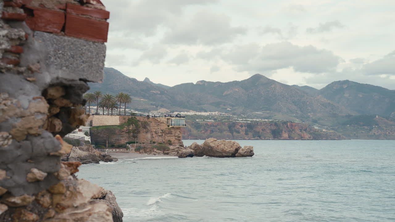 Vista del punto di riferimento Balcon de Europa a Nerja, Spagna, arroccato su spettacolari scogliere costiere sopra il Mar Mediterraneo, con montagne che si ergono sullo sfondo sotto un cielo nuvoloso.