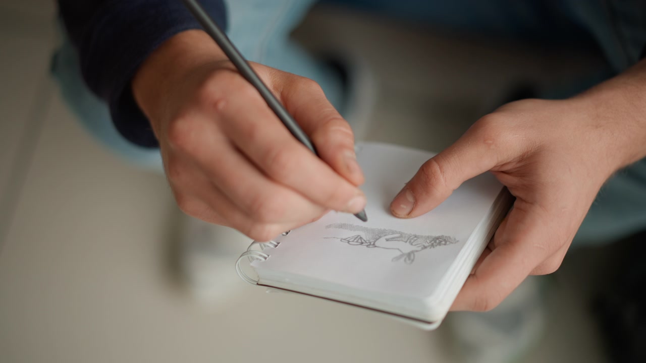 Primo piano di una persona che disegna con una matita su un album da disegno, concentrandosi sul movimento della mano e sull'opera d'arte che emerge. Le pagine del quaderno mostrano leggere linee di grafite mentre il soggetto abbozza una figura animale. Illuminazione interna soffusa.