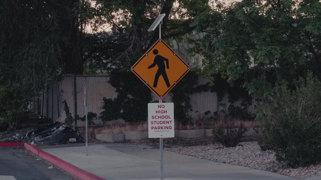 Steady handheld view of a pedestrian crossing sign near a sidewalk lined with trees and landscaping.