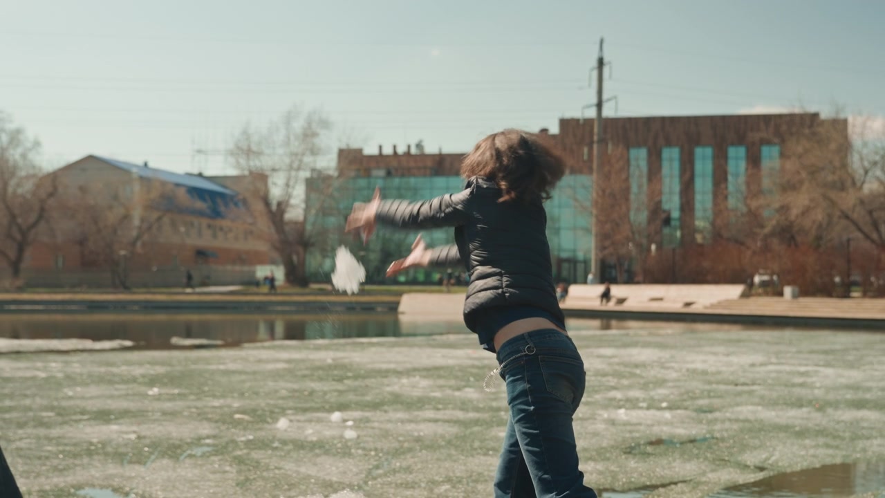 Donna che gioca gioiosamente con il ghiaccio su uno stagno ghiacciato, lanciando e afferrando pezzi mentre indossa abiti invernali casual. La scena presenta un ambiente di parco urbano all'aperto con edifici, alberi e altro.