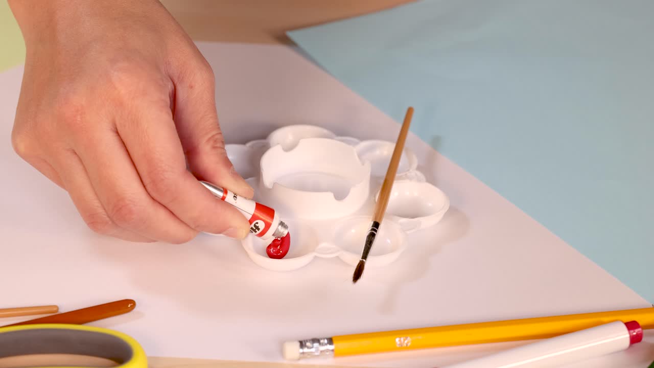 Person dispenses red paint from tube into palette on desk, overhead view, bright lighting