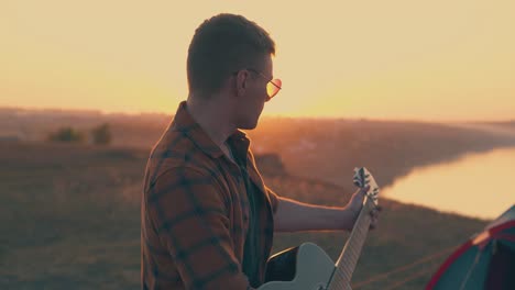 guy plays guitar in camp against river and setting sun