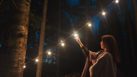 woman caresses string of luminous bulbs. glow from suspended lights adorns trees surrounding camping tent as darkness falls. enchanting atmosphere