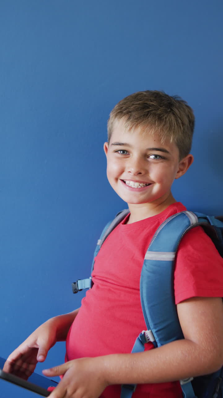 Vertical video of happy caucasian boy wearing backpack on blue background, copy space, slow motion