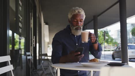 Afroamerikanischer-älterer-Mann,-Der-Sein-Smartphone-Benutzt,-Während-Er-Kaffee-Trinkt-Und-Draußen-Im-Café-Sitzt