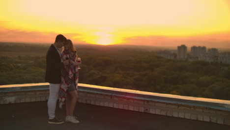 man and young pretty woman sitting on couch holding wine glasses and kissing on rooftop terrace at sunset