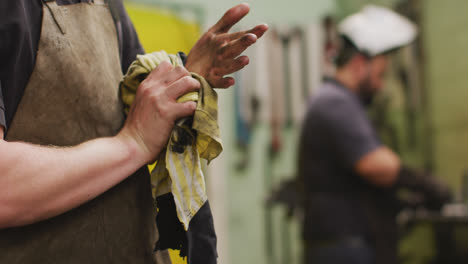 Caucasian-male-factory-worker-at-a-factory-cleaning-his-hands-with-a-cloth,-with-another-man-working