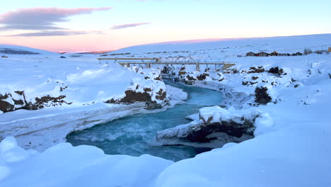 cars drive over the bridges of the godafoss bridges as the meltwater flows quickly between the snowy landscape just after sunset