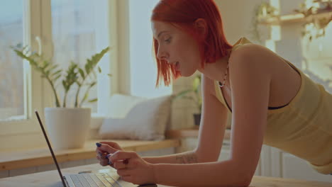 redhead girl leaning on table, typing on laptop in sunny kitchen